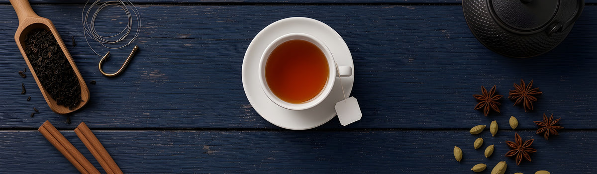 Cup of black tea with a tea bag on a saucer beside a cast-iron teapot, loose tea, a strainer and chai spices on navy dock planks